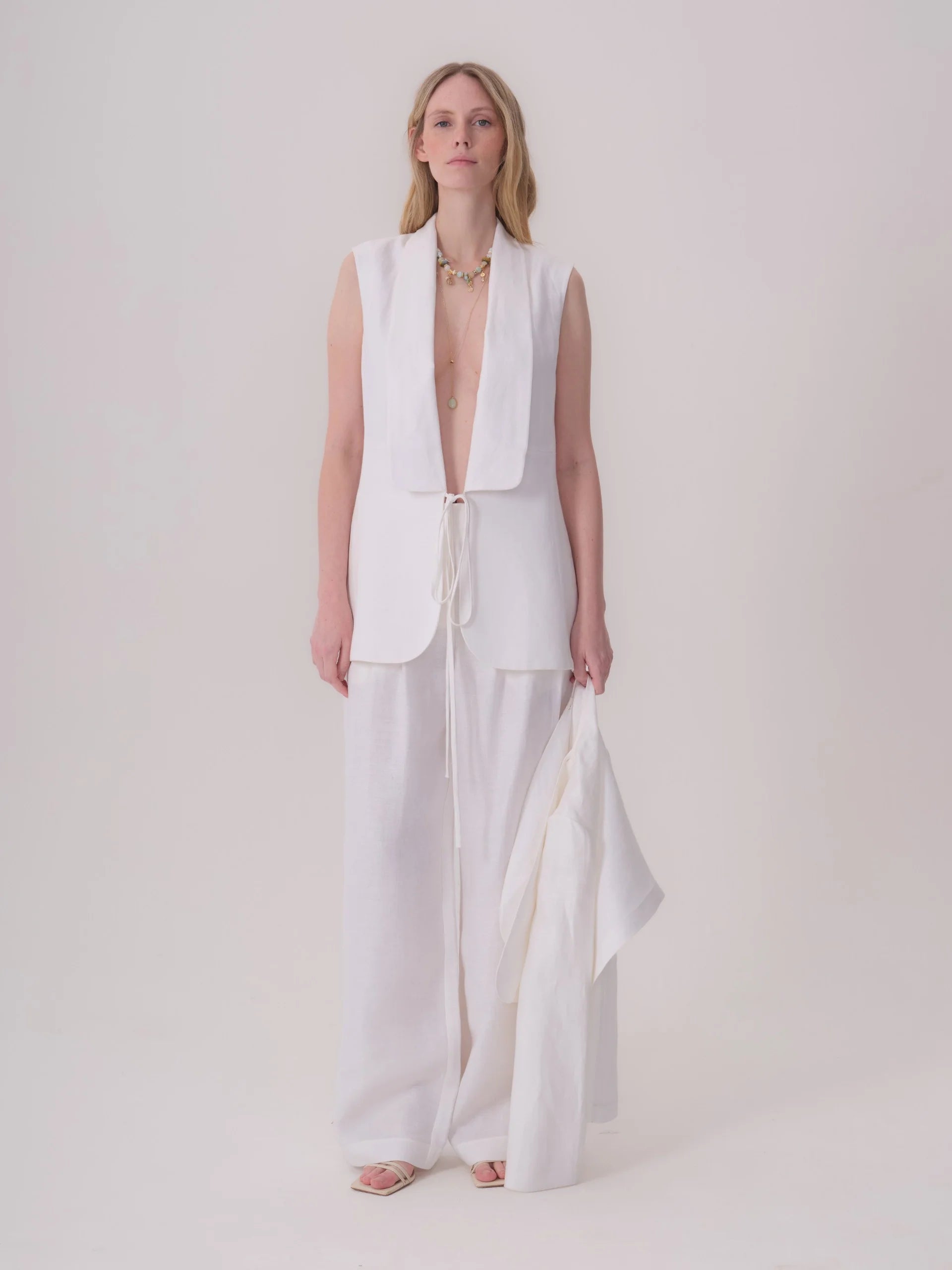 White sleeveless linen-blend jacket with wide shawl lapels and a tie-front closure at the waist.