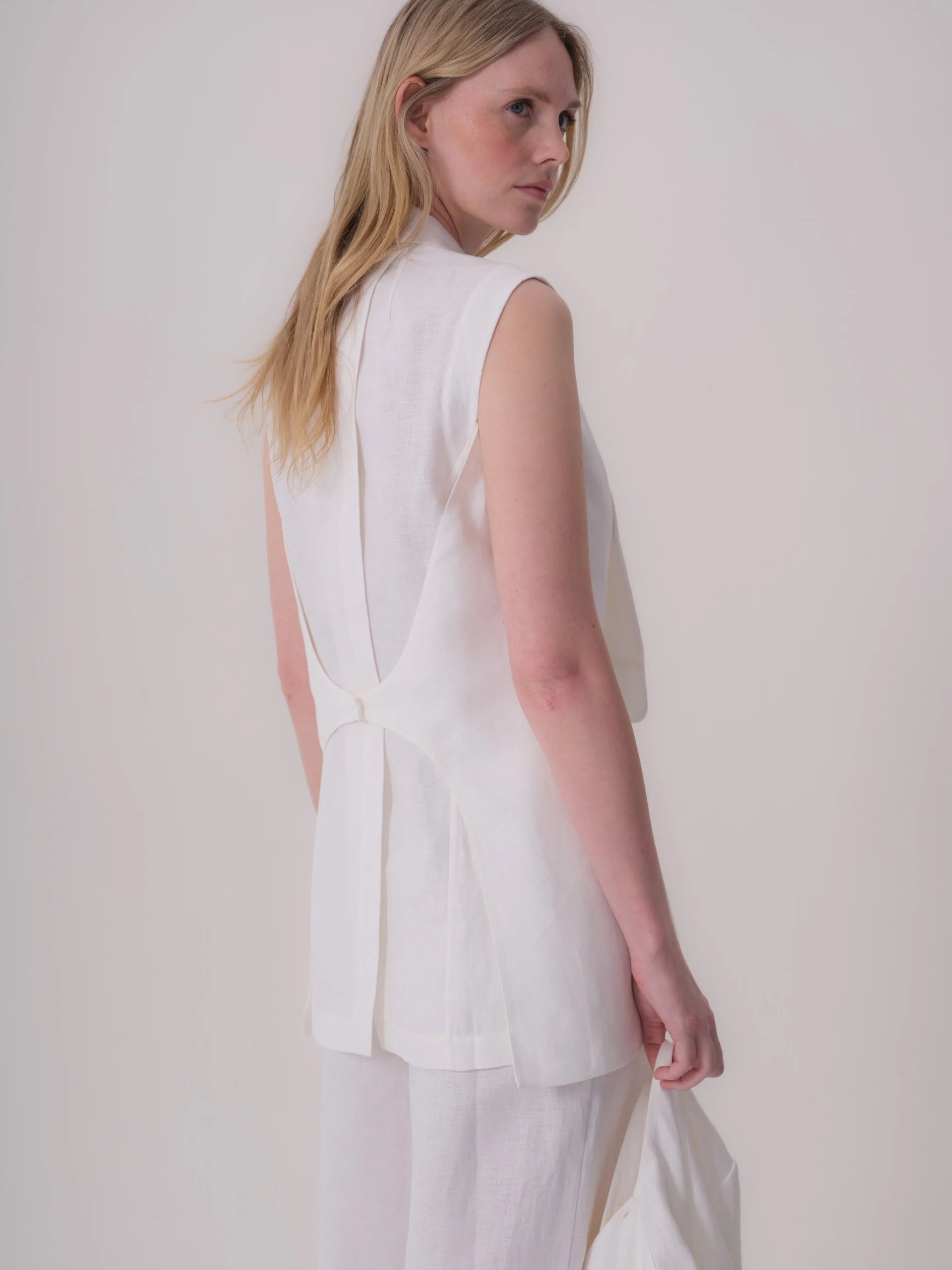 White sleeveless jacket with a stand collar. The back features a central pleat, and a single button closure at the lower back