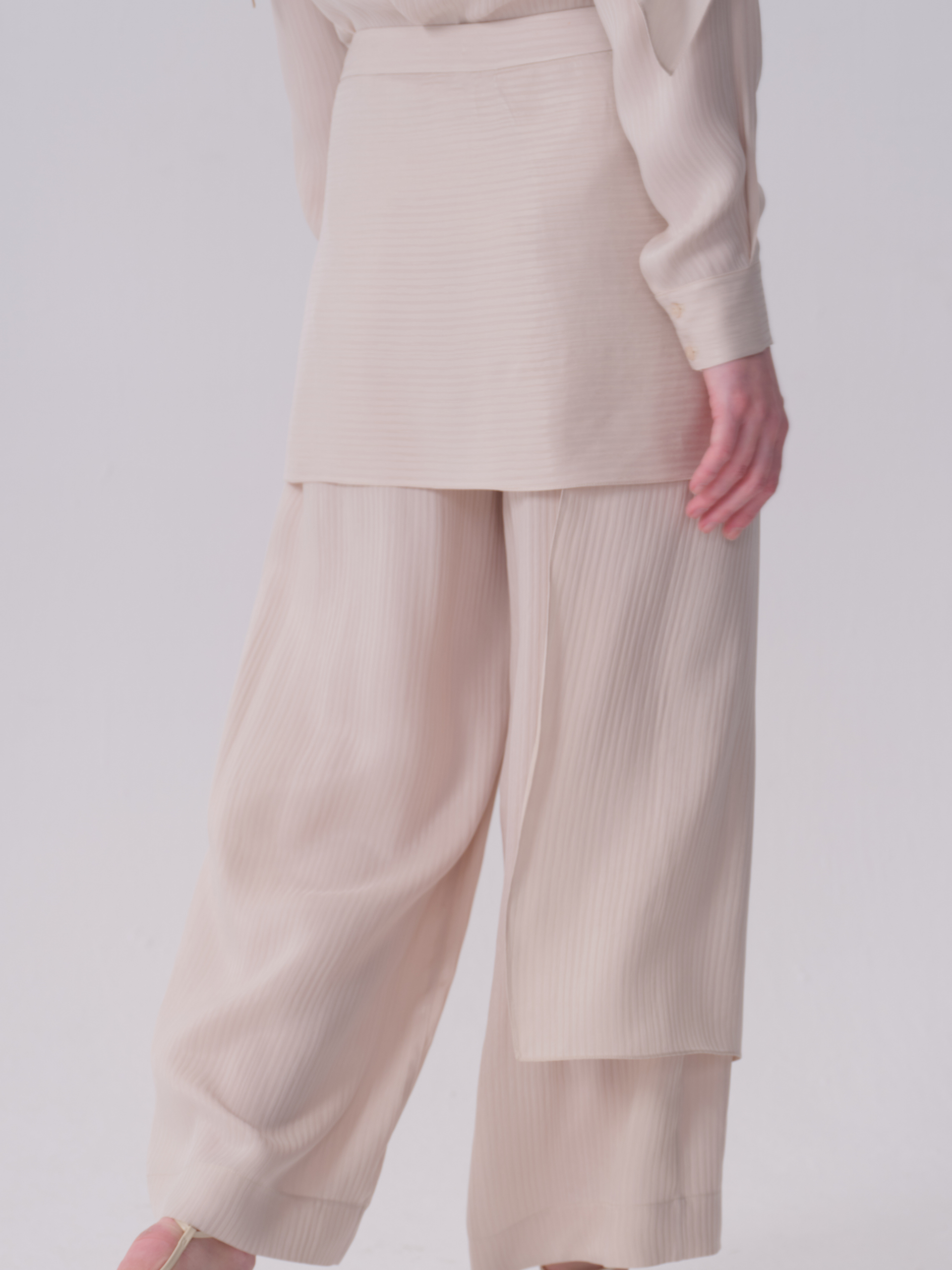 White wide-leg trousers with fine vertical ribbing. The right leg features a layered, wrap-around panel, while the left is a