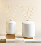White matte ceramic diffuser with a gold-toned band around its rounded top, an embossed logo, and natural reeds.