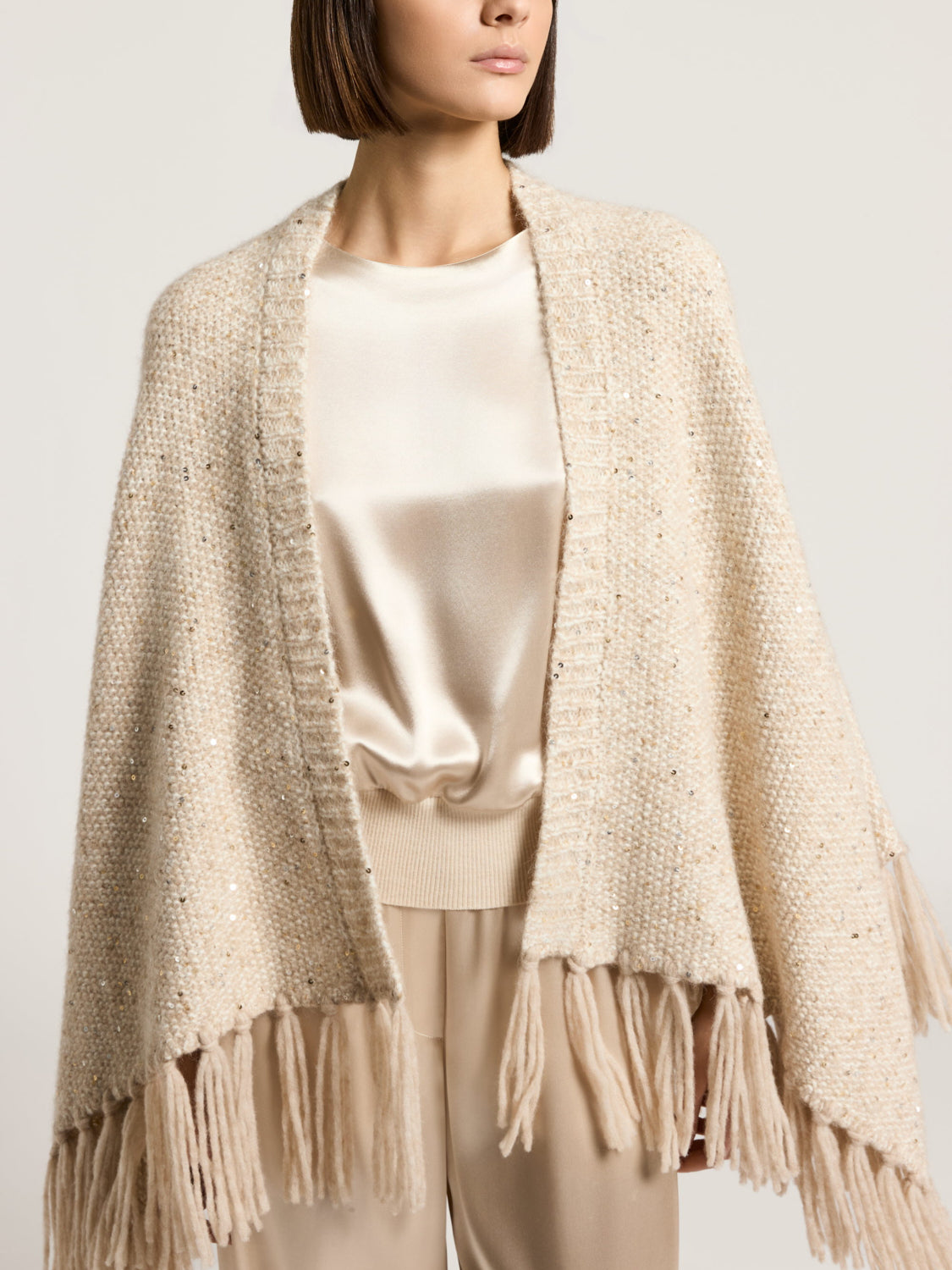 Beige alpaca and wool knit cape with an open front, scattered gold-toned sequins, and fringe along the bottom edge.