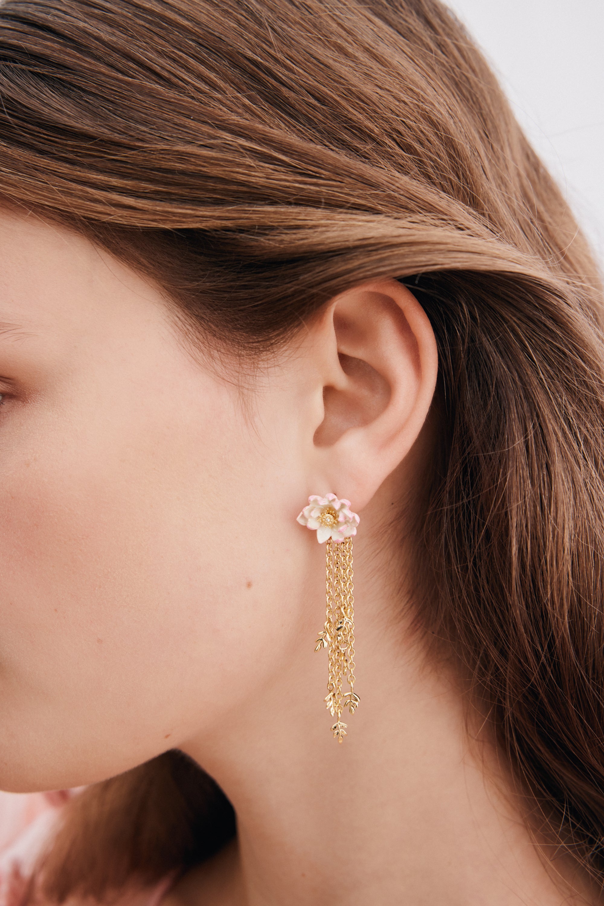 A single post earring with a light pink and white flower top, yellow center. Gold-toned chains dangle with small leaf charms.