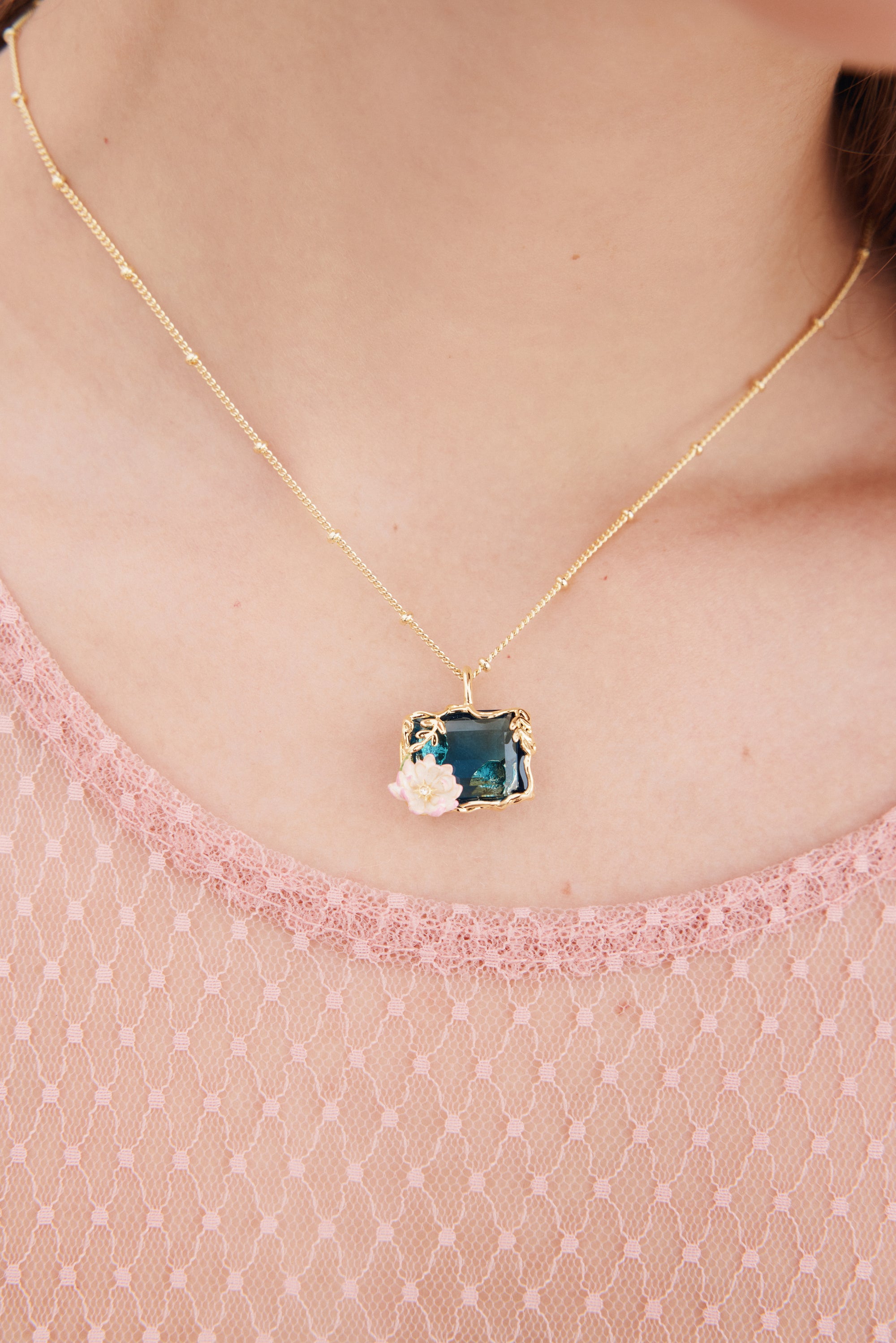 Gold-toned beaded chain necklace with a rectangular dark teal pendant framed by gold-toned leaves and pink and white flowers.