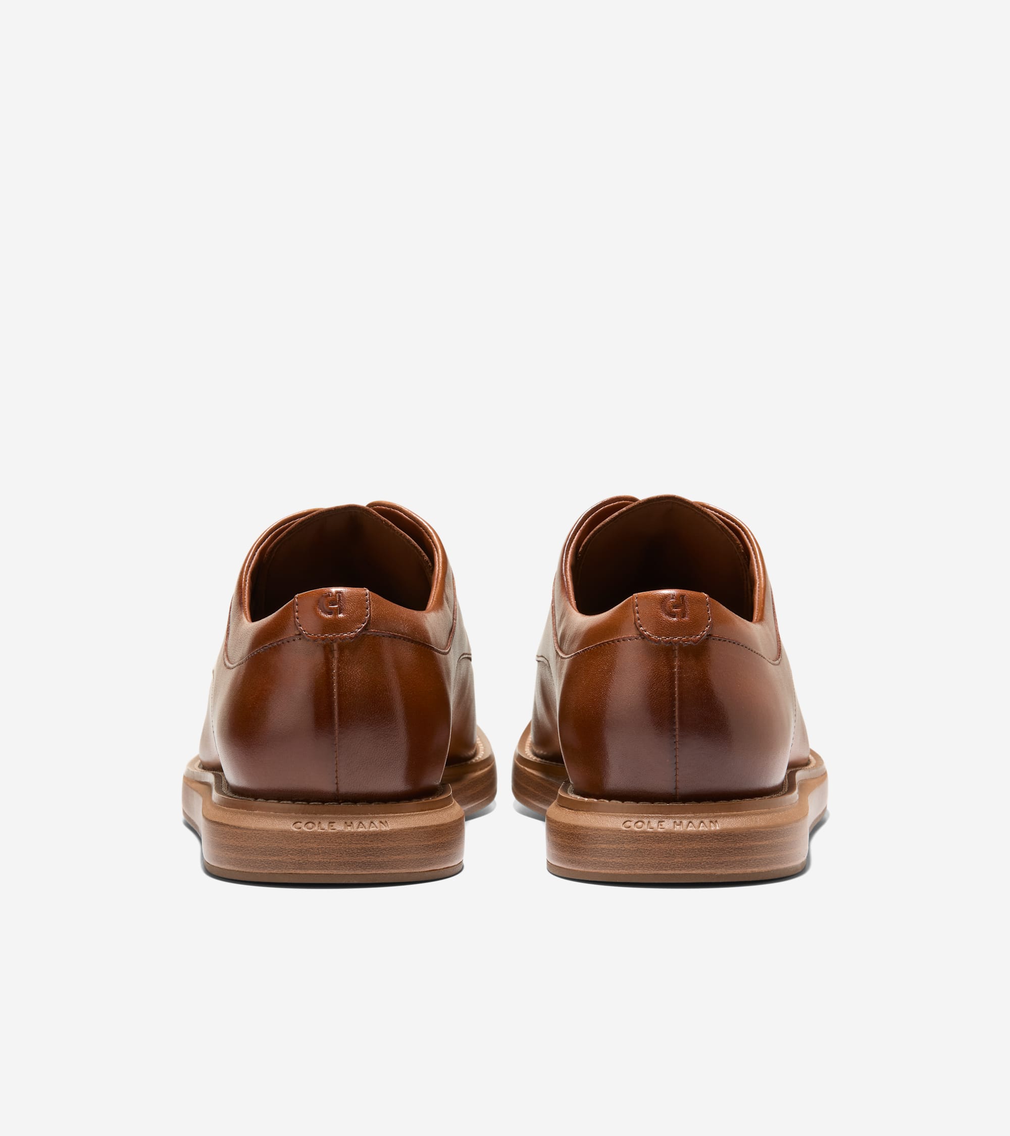 Pair of polished brown leather oxfords, seen from the back. Each shoe has an embossed 'CH' logo on the heel and 'COLE HAAN' o