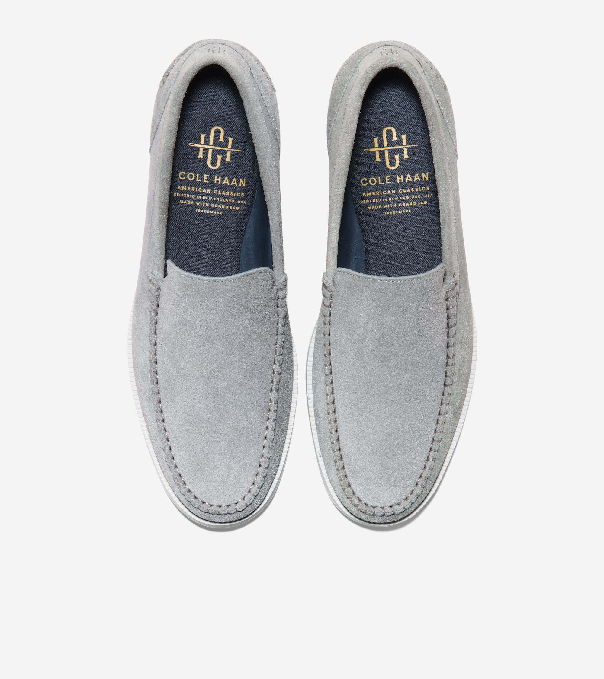 A pair of light gray suede loafers with moc-toe stitching, white rubber soles, and dark blue insoles with gold-toned Cole Haa
