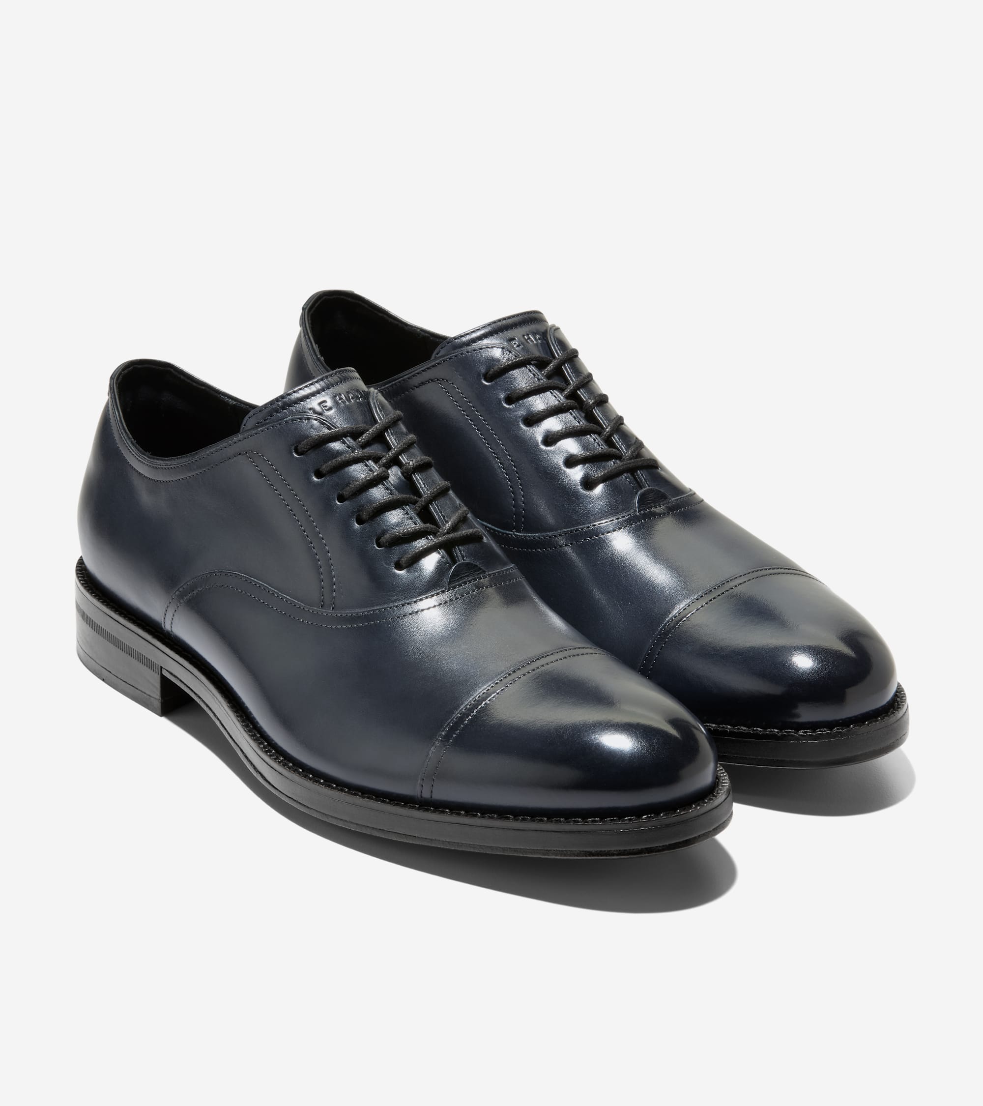 A pair of dark blue polished leather cap toe oxfords with black laces, black soles, and visible stitching.