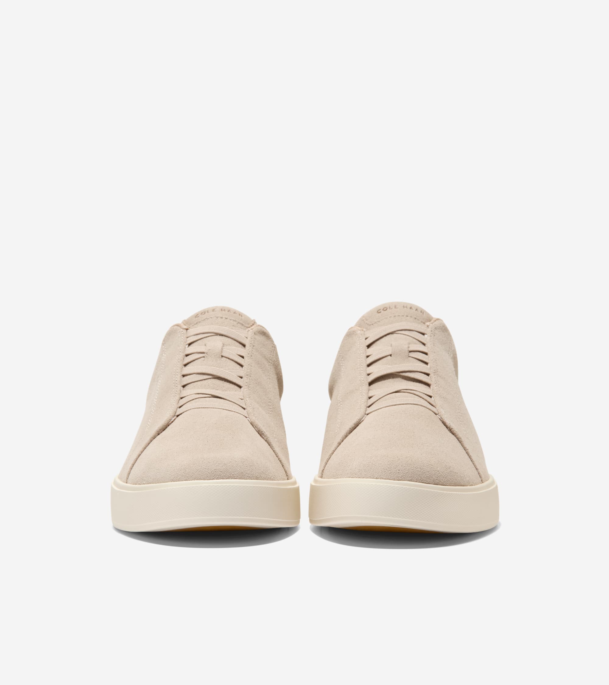 Pair of sand-colored suede slip-on sneakers with decorative elastic bands, thick cream soles, and 'COLE HAAN' on the tongue.