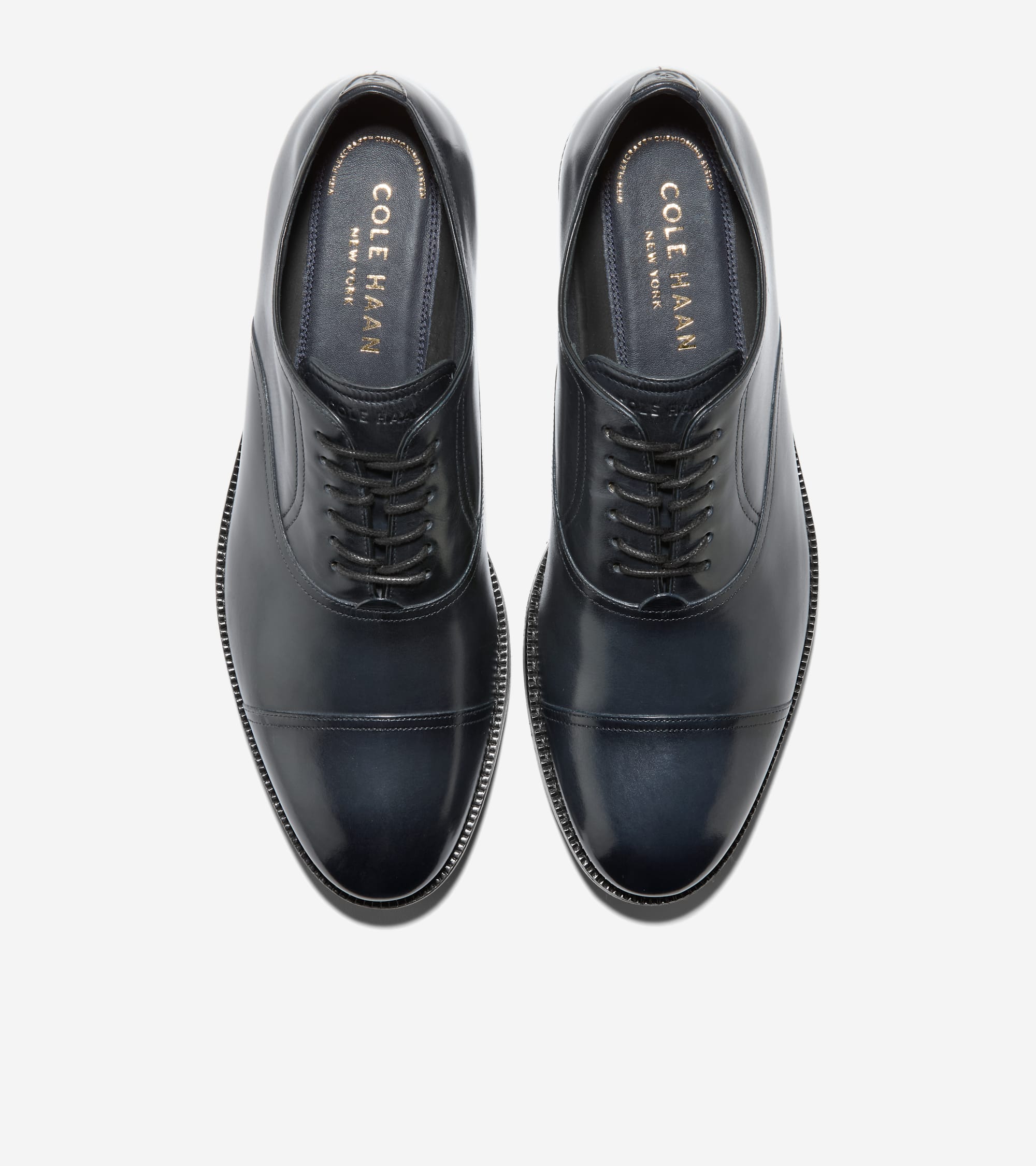 A pair of dark blue polished leather cap toe oxford shoes with dark laces and gold-toned Cole Haan logo on the insoles.