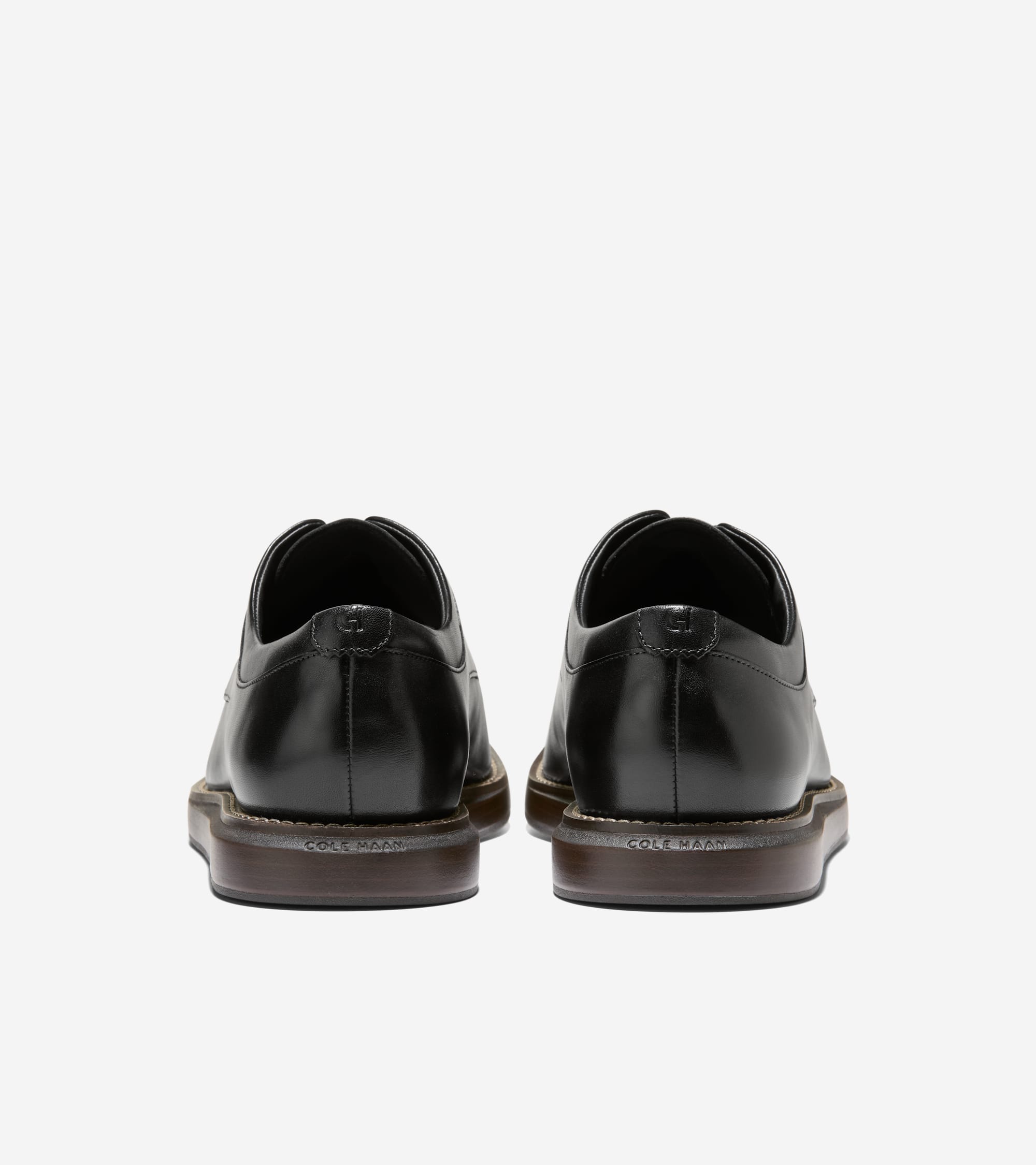 A pair of black polished leather oxfords, seen from the back, with dark brown soles. "COLE HAAN" is on the sole, and a "CH" l