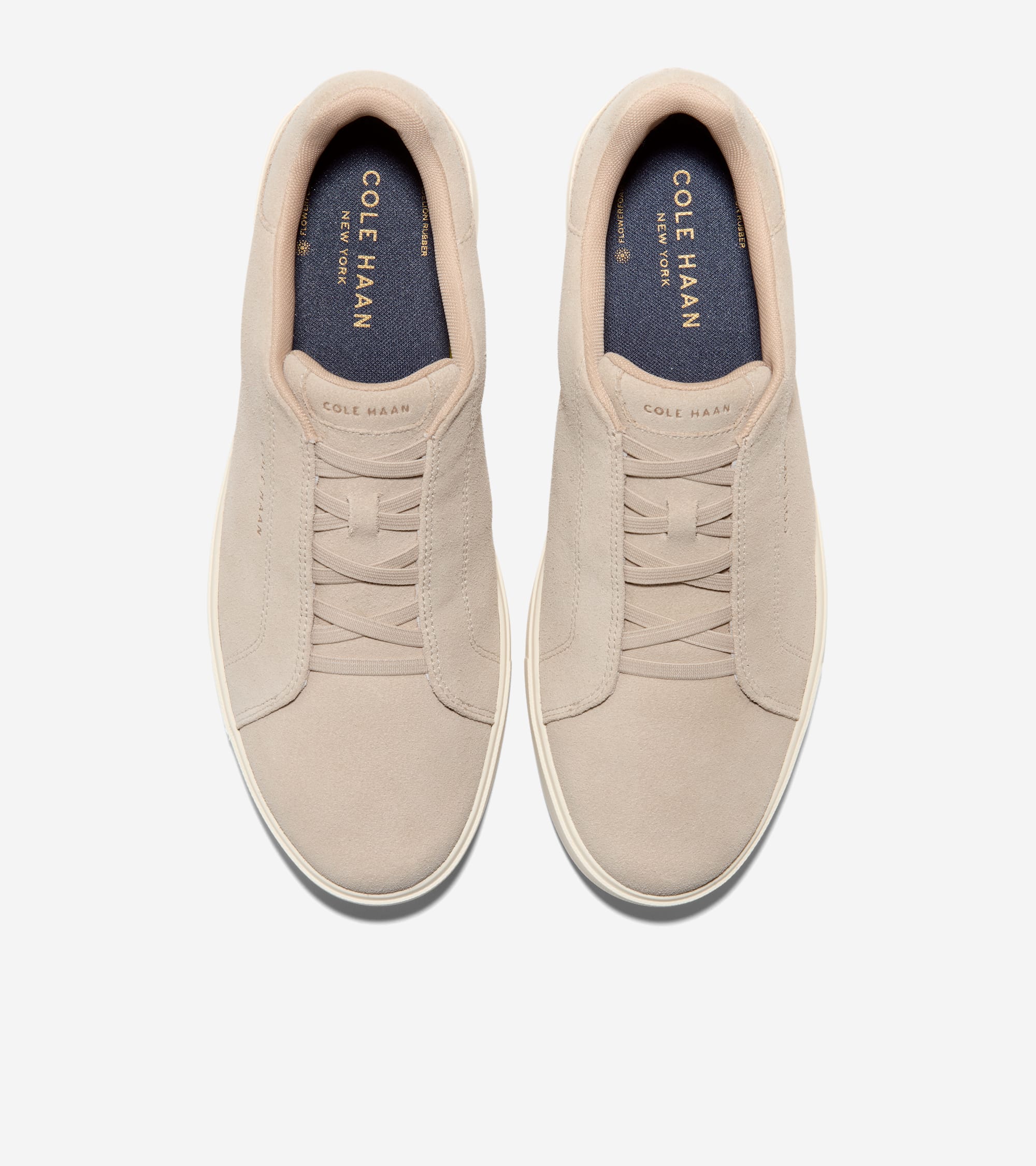 Pair of light beige suede slip-on sneakers with elastic criss-cross bands on the vamp and white soles. Dark blue footbeds.