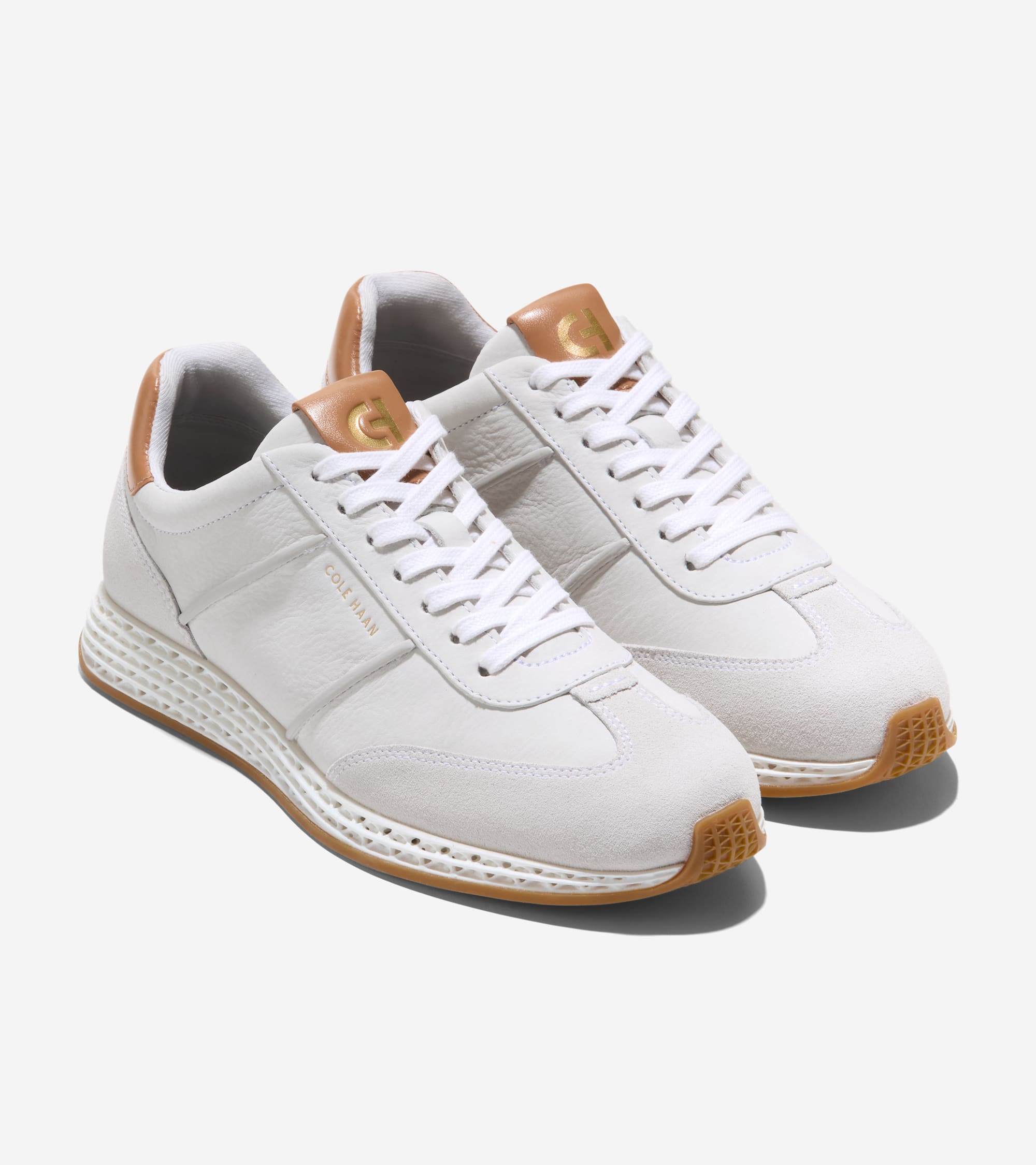 Off-white sneakers with textured uppers, suede toe caps, white laces, tan heel/tongue accents with gold-toned logos, and gum