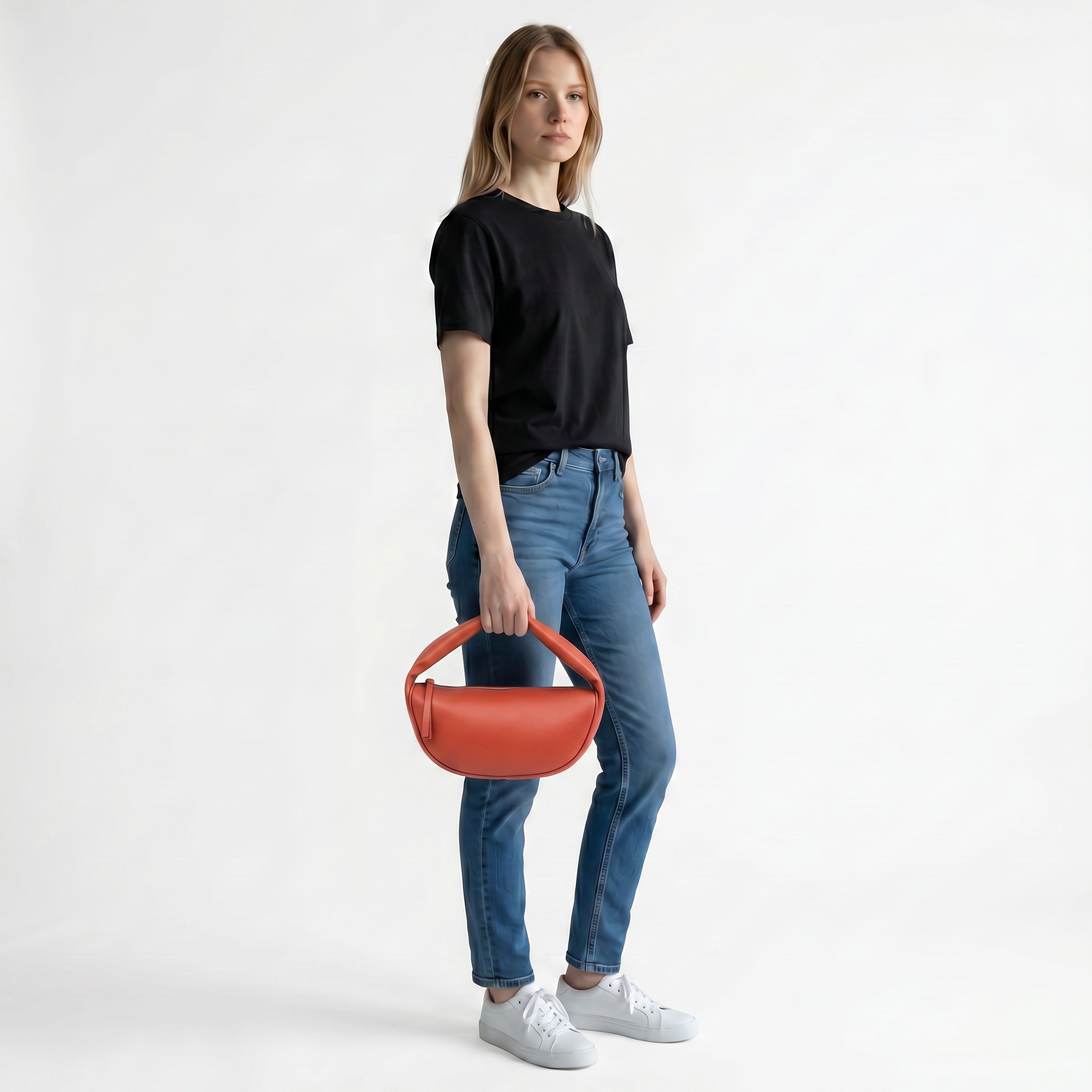A model wearing a black t-shirt and blue jeans holds the burnt orange By Far crescent leather handbag to demonstrate its scale, from BLUE SALON.