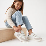 White low-cut sneakers with light grey toe and side panels, three white velcro straps, a white 'CK' logo, and black heel tab.