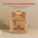 Fluffy beige teddy backpack with rounded ears, a dark zippered front pocket, and black 'MY FIRST BAG' text. Brown top handle.