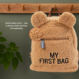 Beige teddy fleece backpack with rounded ears, a front zipper pocket with a gray "CHILDHOME" pull, and "MY FIRST BAG" embroid
