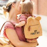 Beige teddy bear-style backpack with two rounded ears, a front zipper pocket, and 'MY FIRST BAG' embroidered in black.