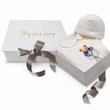 White sleepsuit and hat set. Sleepsuit features a teddy bear in a blue jacket peeking from a pocket, holding a gold-toned poc