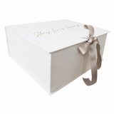 White rectangular gift box with a hinged lid, gold-toned 'Story Loves Luxury' script, and a light taupe satin ribbon bow.