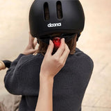 Black helmet with two vertical vents, white 'doona' logo, and a glowing red oval light on the back adjustment strap.