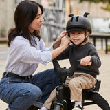 Black kids' helmet with a small brim, ventilation slots, and two horn-like protrusions on top. A chin strap is buckled.