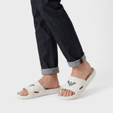 Light beige rubber slide slippers with a wide strap, featuring a black circular logo on each strap and textured footbeds.