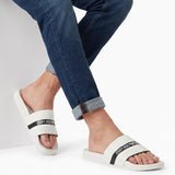 Pair of white slide sandals with a wide, ridged strap. A black stripe with "EMPORIO" and an eagle logo in white crosses the s