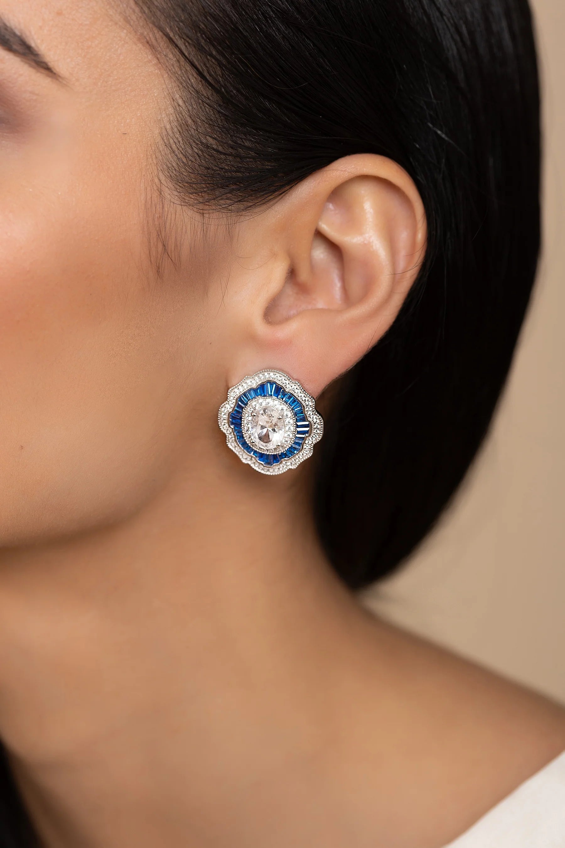 Silver-toned Art Deco stud earring with a central oval clear stone, surrounded by blue baguette stones and a scalloped border