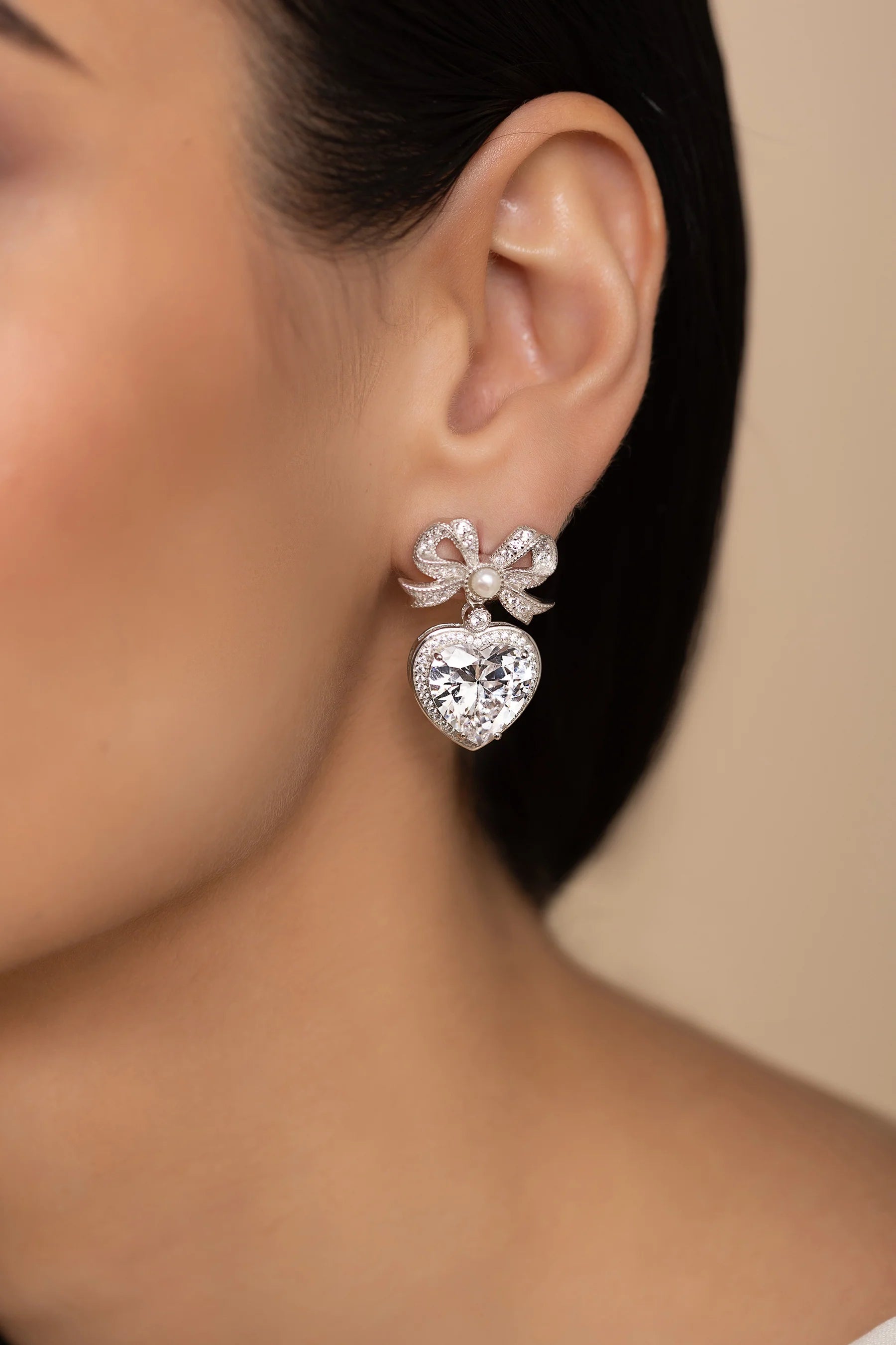 Silver-toned bow earring with clear crystals and a pearl, dangling a large clear heart-shaped crystal framed by smaller cryst
