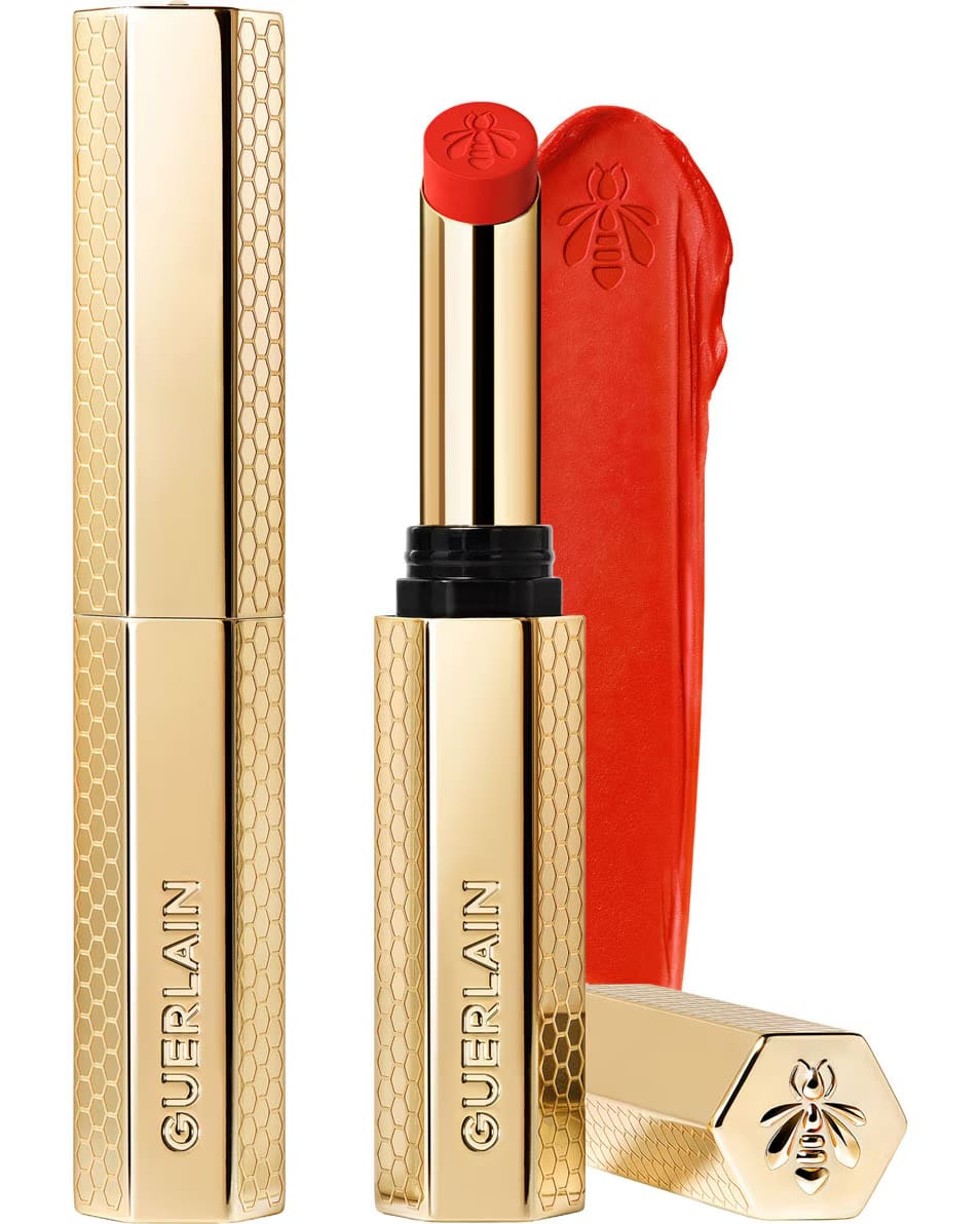 Guerlain KissKiss Honey-Infused Matte Lipstick in its signature slim golden honeycomb case, alongside an open vibrant coral-red lipstick bullet and creamy velvety texture smudge, available at blue salon.