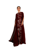 A woman modeling a Velvety brand maroon evening gown with floral lace and sequin embroidery. The dress features a round neckline and a sheer, floor-length chiffon cape attached to the shoulders, set against a textured sunlit wall.