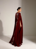 A woman modeling a Velvety brand maroon evening gown with floral lace and sequin embroidery. The dress features a round neckline and a sheer, floor-length chiffon cape attached to the shoulders, set against a textured sunlit wall.