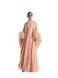 A woman modeling a Velvety brand peach-colored chiffon gown. The dress has a boat neckline, a loose-fitting kaftan silhouette, and dramatic sleeves finished with thick, matching peach ostrich feathers.