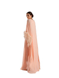 A woman modeling a Velvety brand peach-colored chiffon gown. The dress has a boat neckline, a loose-fitting kaftan silhouette, and dramatic sleeves finished with thick, matching peach ostrich feathers.