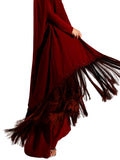 Side view of woman modeling a Velvety brand burgundy two-piece set consisting of a sleek square-neck column dress and a matching long-sleeved sheer outer cape with heavy fringe detailing at the hem.