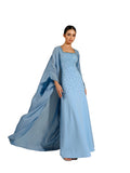 A woman modeling a Velvety brand sky-blue two-piece set. The inner dress is a column-style gown with a square neckline and extensive tonal beadwork. The outer layer is a matching floor-length cape with a crinkled texture and wide, flowing sleeves.