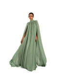 A woman modeling a Velvety brand evening gown in green. The dress features a high-neck halter design, tonal bead embellishments on the chest, and a voluminous floor-length cape attached to the shoulders.