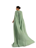 Back view of a woman modeling a Velvety brand evening gown in green. The dress features a high-neck halter design, tonal bead embellishments on the chest, and a voluminous floor-length cape attached to the shoulders.