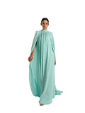 A woman modeling a Velvety brand evening gown in tifany. The dress features a high-neck halter design, tonal bead embellishments on the chest, and a voluminous floor-length cape attached to the shoulders.