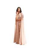 A woman modeling a Velvety brand nude evening gown with floral lace and sequin embroidery. The dress features a round neckline and a sheer, floor-length chiffon cape attached to the shoulders, set against a textured sunlit wall.
