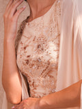 A woman modeling a Velvety brand nude evening gown with floral lace and sequin embroidery. The dress features a round neckline and a sheer, floor-length chiffon cape attached to the shoulders, set against a textured sunlit wall.