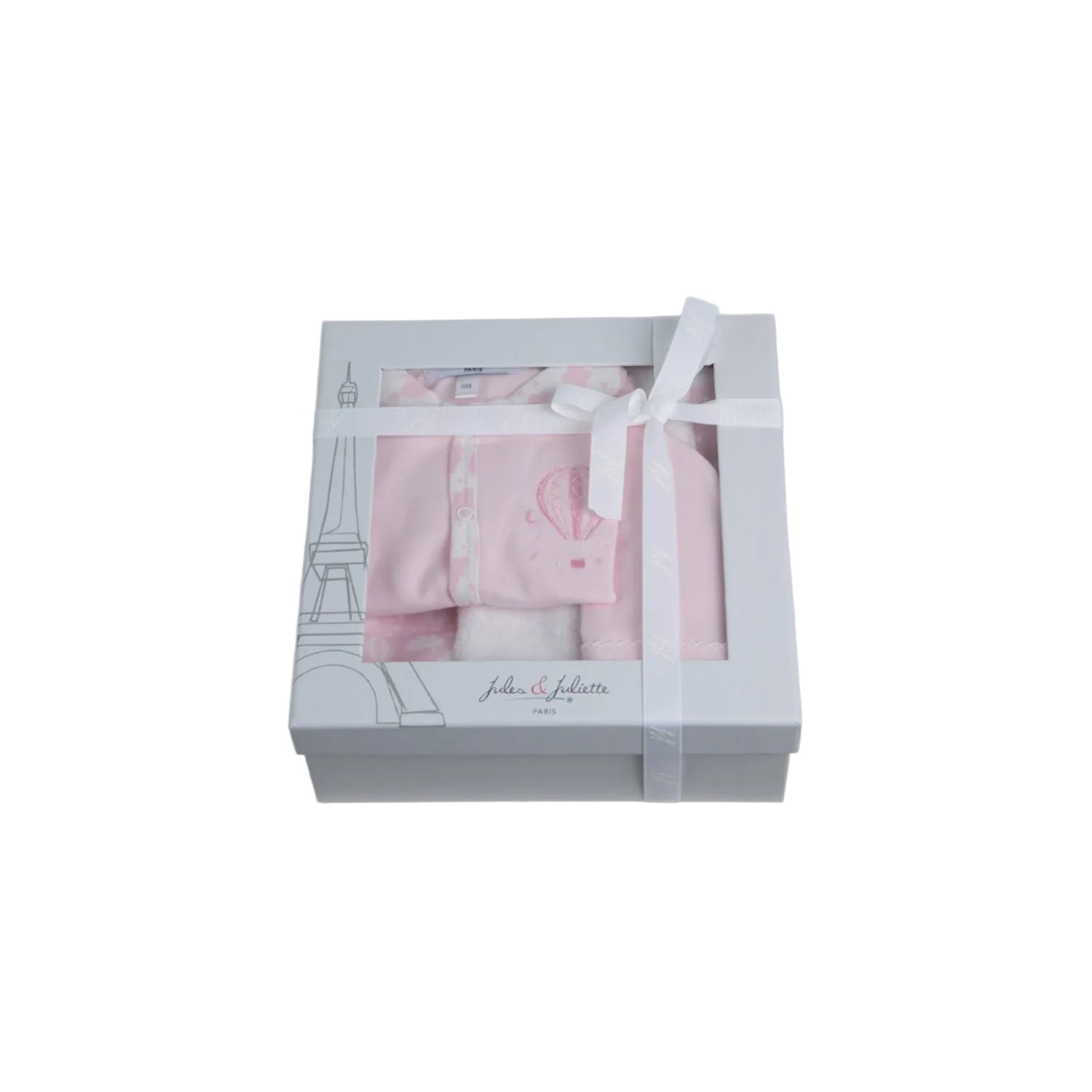 A luxury 3-piece baby girl gift set by Jules & Juliette Paris in light pink. Images show a footed romper with hot air balloon embroidery, a matching pink beanie with picot trim, and a reversible white and pink patterned blanket. The set is also shown packaged in a signature grey window gift box with a white ribbon.