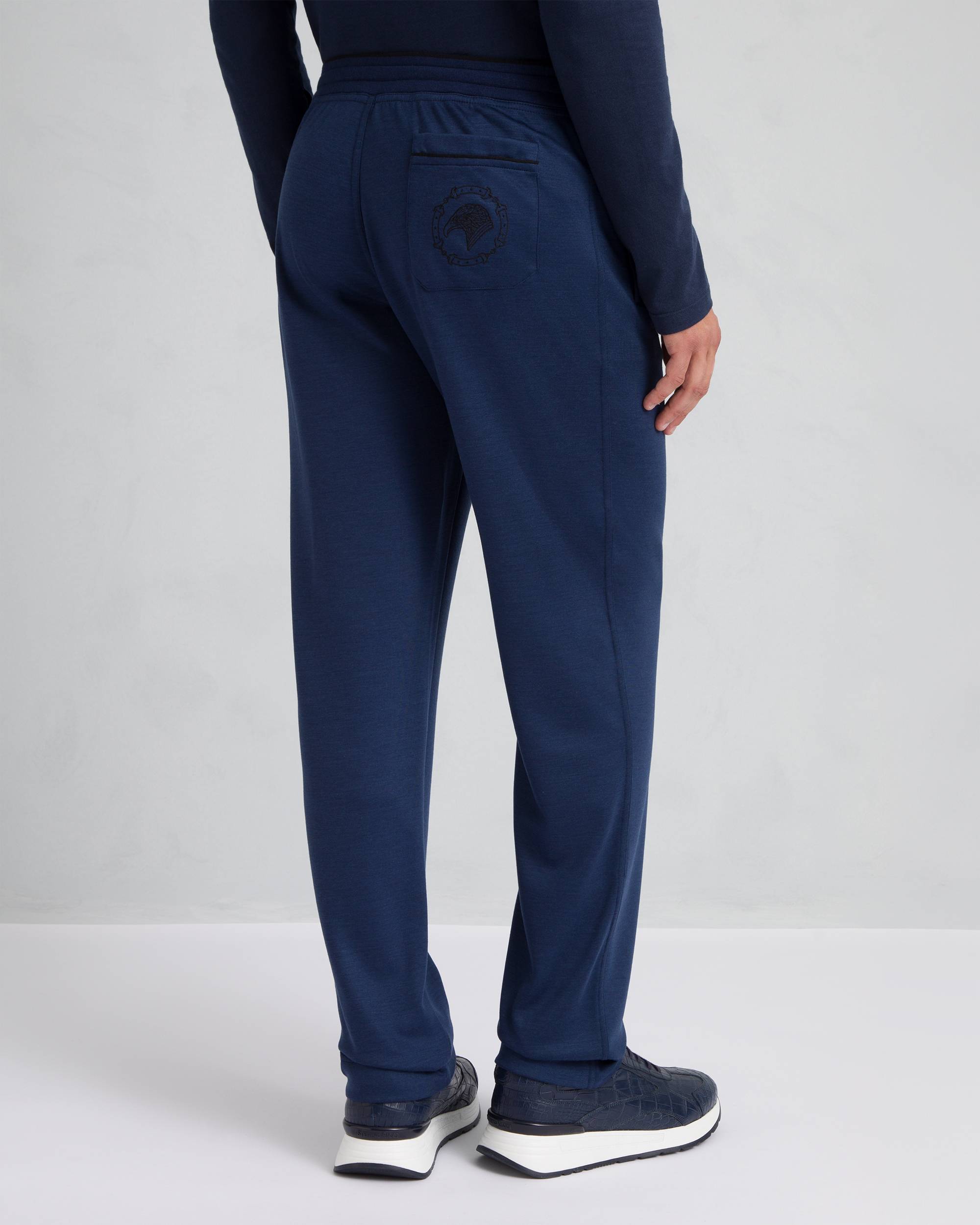 Blue jersey knit trousers with an elasticated drawstring waist, a back pocket with a dark circular embroidered logo, and stra