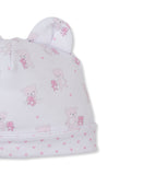 White baby hat with two rounded ears, patterned with pink teddy bears (some holding smaller bears) and scattered pink hearts.