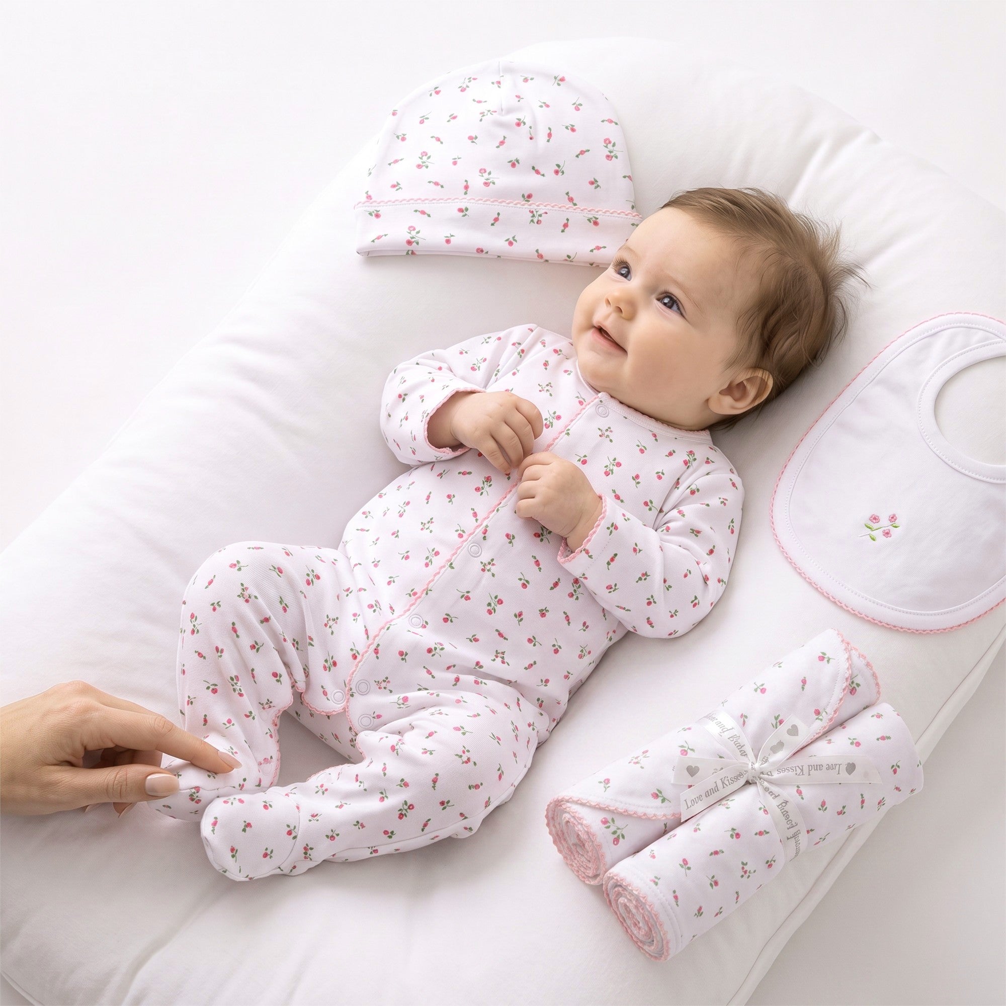 White baby gift set: floral print footie sleeper, matching hat, white bib with pink rose embroidery, and two rolled floral bl