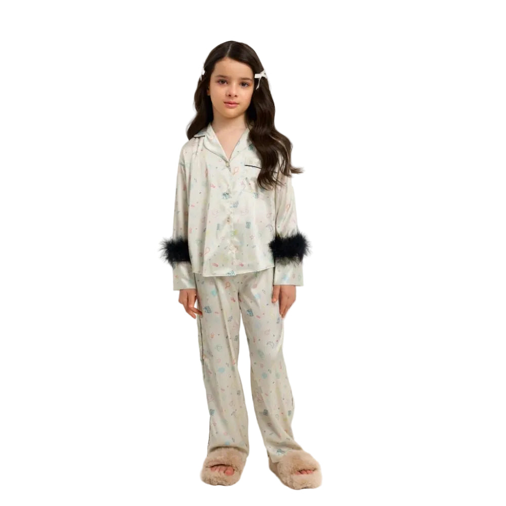A professional studio shot of a young girl wearing a Katee's Kids ivory silk pajama set featuring a colorful star and moon print, highlighted by dramatic black faux fur trim on the sleeves, set against a solid white background.