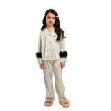 A professional studio shot of a young girl wearing a Katee's Kids ivory silk pajama set featuring a colorful star and moon print, highlighted by dramatic black faux fur trim on the sleeves, set against a solid white background.