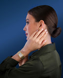 Side profile lifestyle view of a model resting her hand against her face, elegantly showcasing the Saint Honoré Palais Royal gold watch on her wrist against a rich blue background.