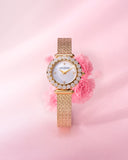 A lifestyle studio shot of the Saint Honoré Gala 29mm watch nested in front of soft pink carnations, highlighting the gold-plated mesh band and the pavé diamond flower-motif bezel.