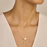 Gold-toned necklace with a thin chain. Pendant features a textured, wavy gold-toned bar with an irregular white pearl and sma