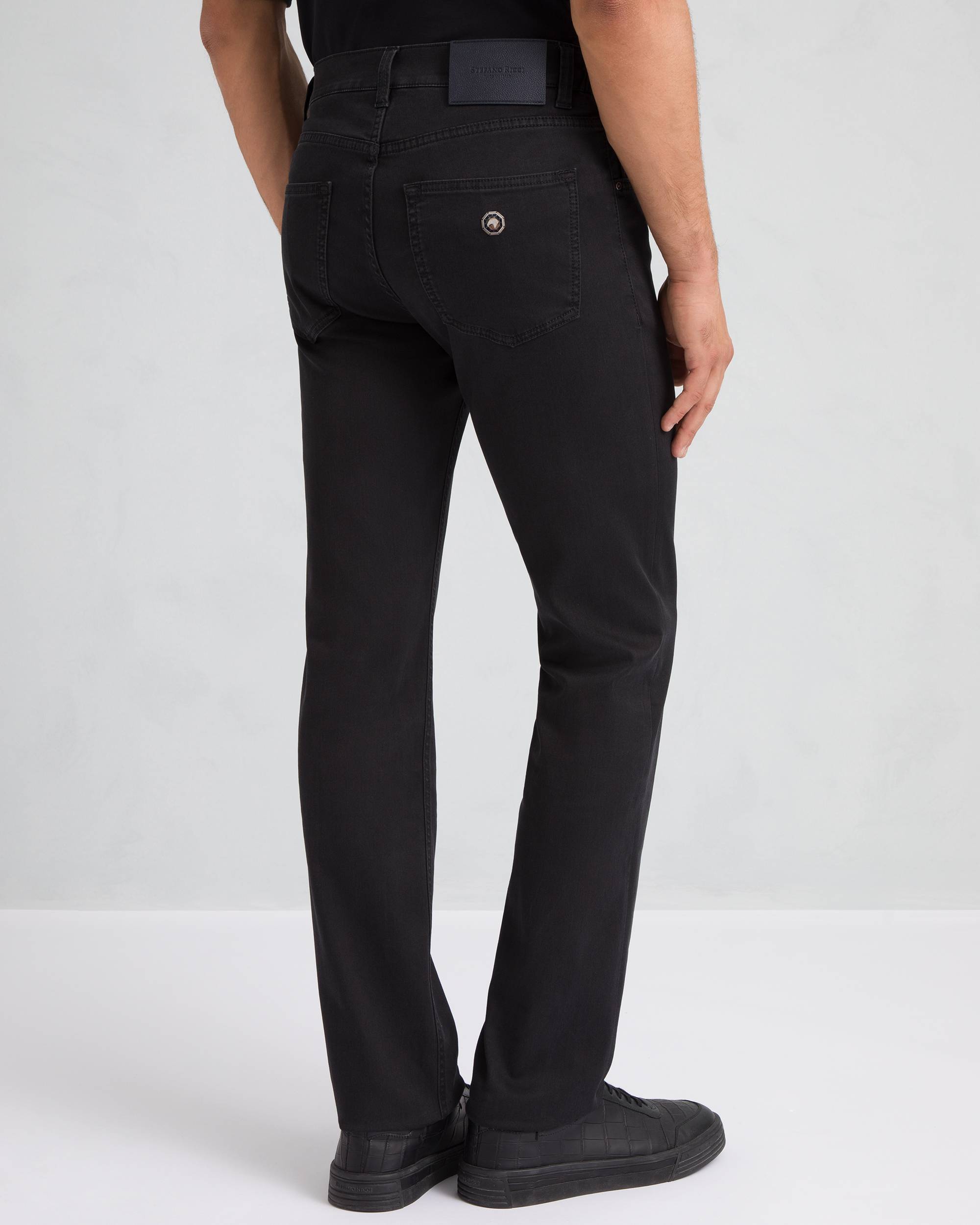 Black dark wash trousers with a black brand patch on the back waistband and a silver-toned circular logo button on the right